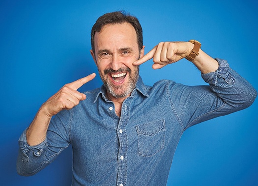 An older man wearing a denim shirt and pointing to his new smile thanks to All-On-4 implants in Uptown New Orleans